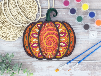 Finished mandala pumpkin adult craft kit shown with painted layered wood artwork, unpainted kit pieces, paint pots, and brushes on a rustic wood background.