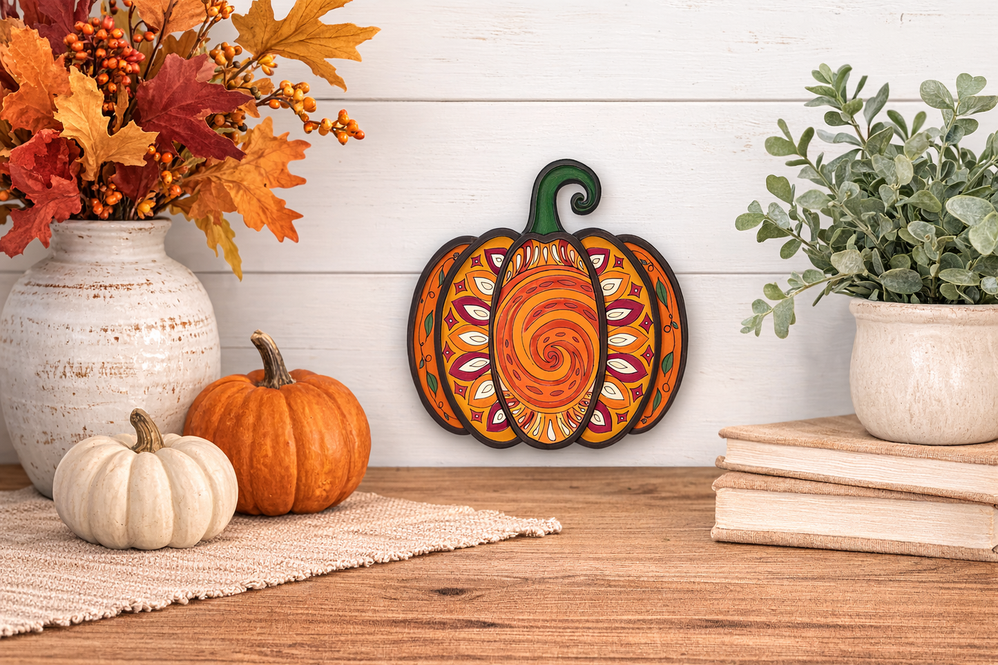 Completed layered wood paint kit completed and shaped like a pumpkin with a mandala design hanging on a wall above a wooden surface with pumpkins and plants.