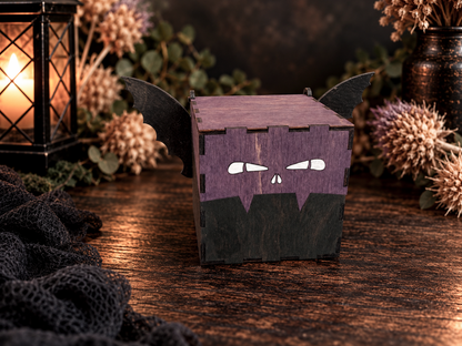 Purple handmade wooden bat keepsake box with black wings, hand-painted facial details, and hinged lid surrounded by dried flowers and warm lantern light.