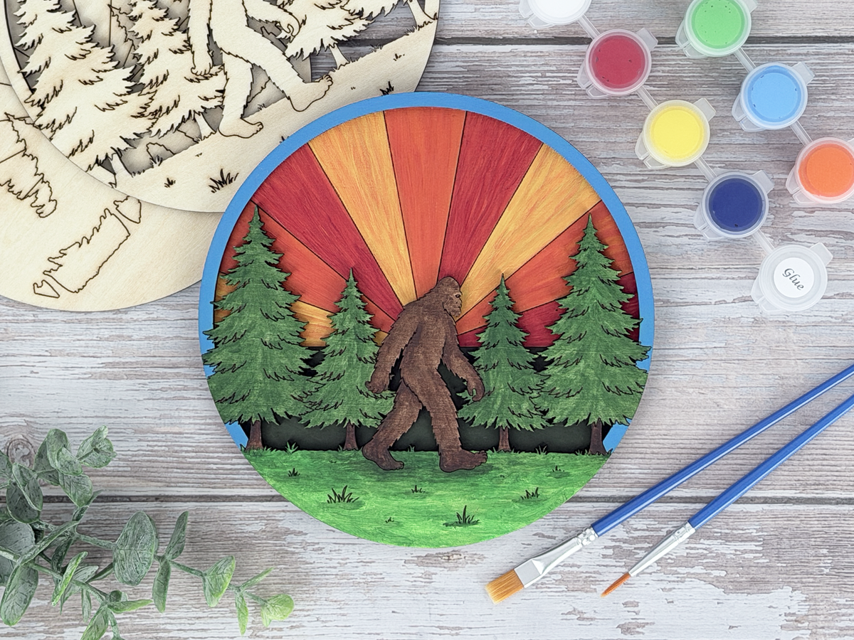 Finished sasquatch adult craft kit shown with painted layered wood artwork, unpainted kit pieces, paint pots, and brushes on a rustic wood background.