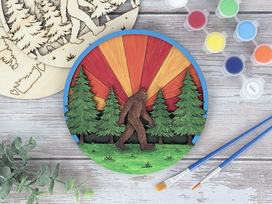 Finished sasquatch adult craft kit shown with painted layered wood artwork, unpainted kit pieces, paint pots, and brushes on a rustic wood background.