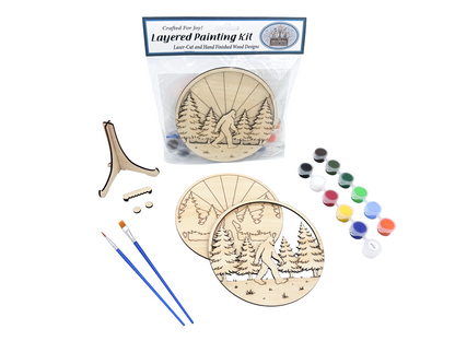 Packaged Sasquatch adult craft kit with unpainted layered wood pieces, paint pots, brushes, display stand, and hardware on a white background.