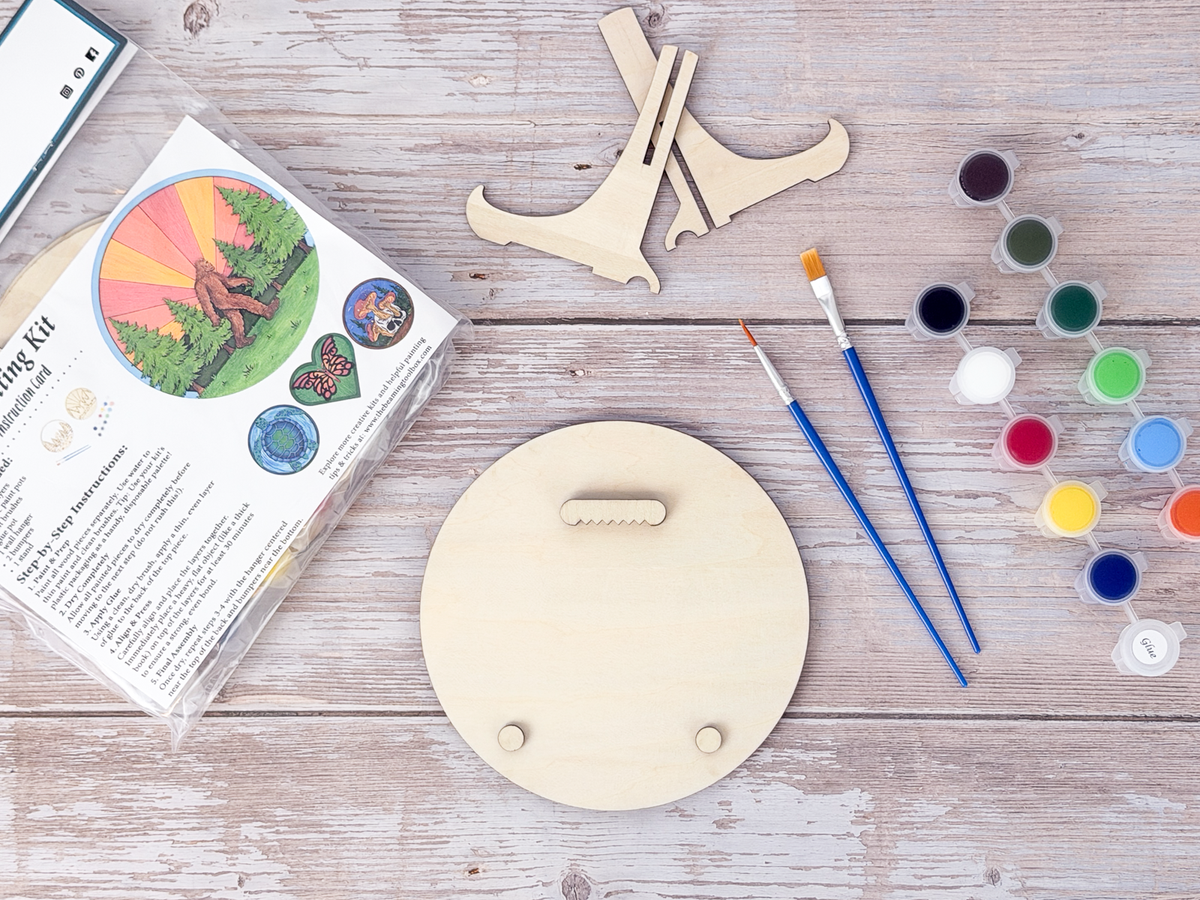 Back view of a Sasquatch adult craft kit showing the wood hanger piece, display stand parts, paints, brushes, and packaged kit on a rustic wood background.