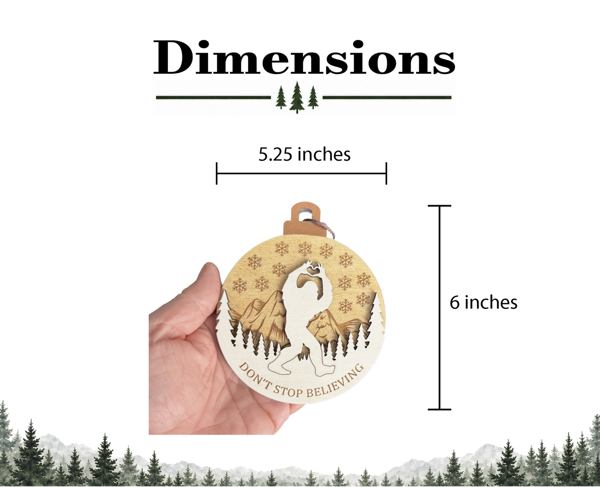 A round wooden ornament featuring a cutout of a Bigfoot making a heart shape with its hands, set against a mountain and forest background with snowflakes. The text reads DON'T STOP BELIEVING. It measures 5.25 inches wide by 6 inches high.
