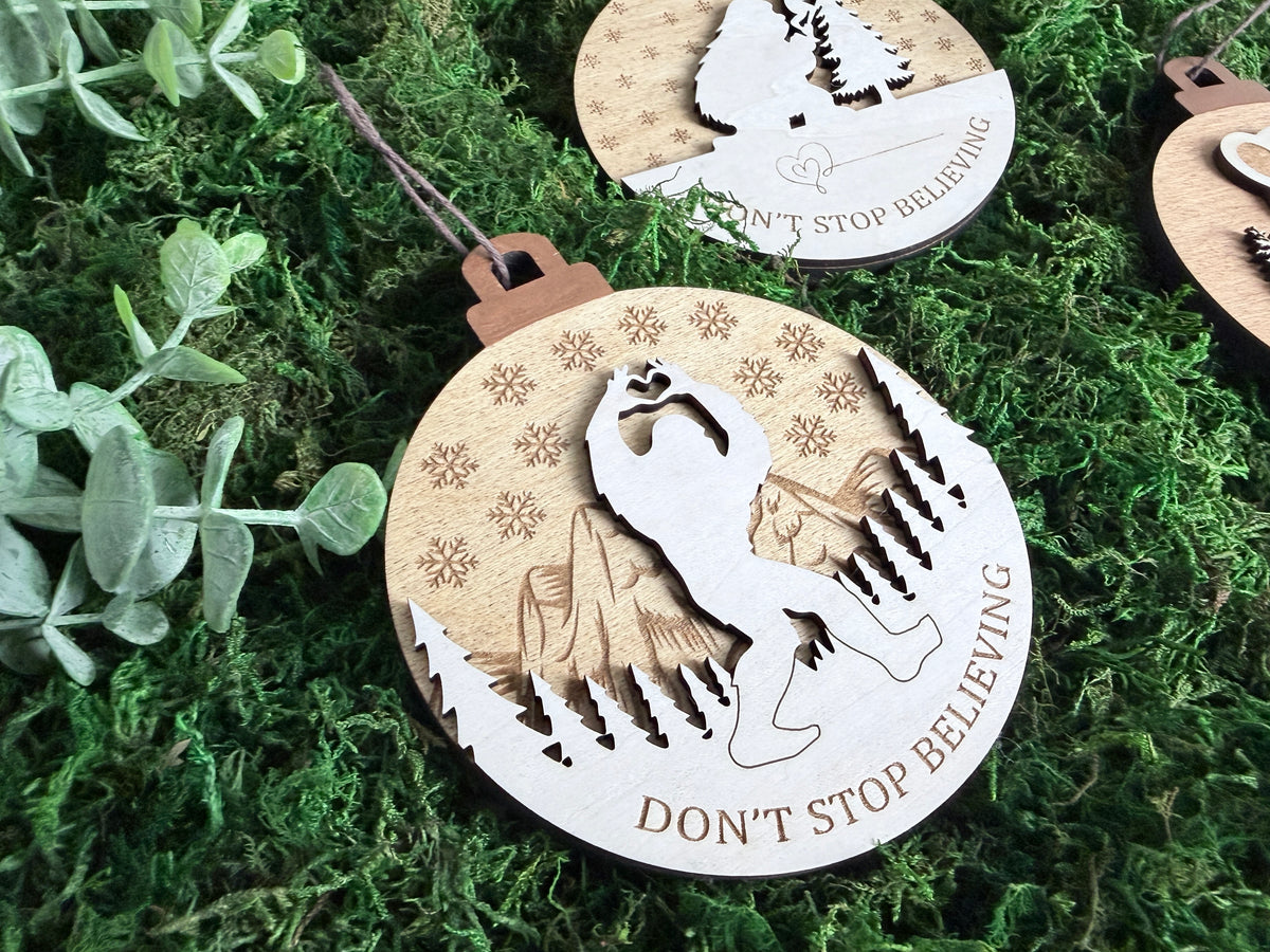 Close-up of layered wooden Bigfoot Christmas ornament featuring a Sasquatch silhouette, engraved mountains, pine trees, snowflakes, and Don't Stop Believing.