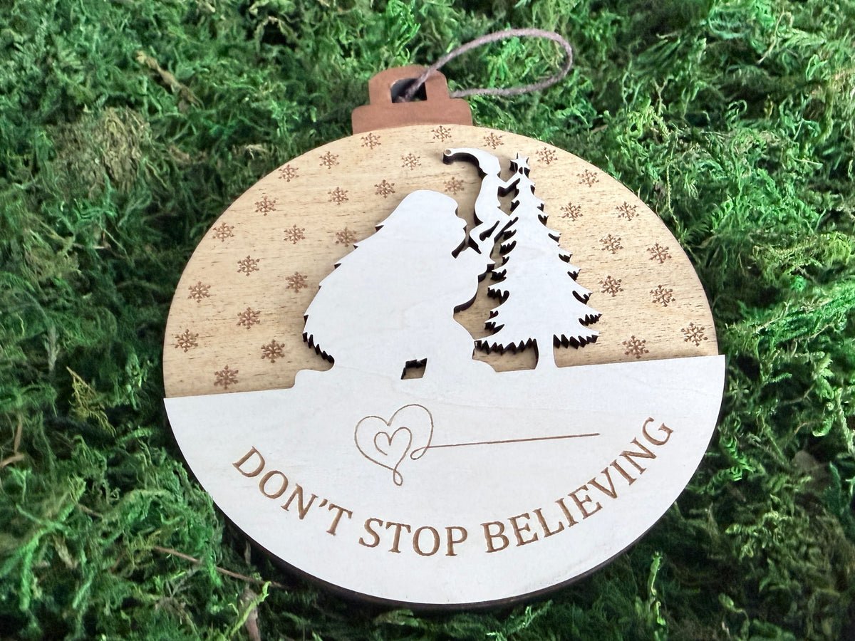 Layered wooden Sasquatch Christmas ornament showing Bigfoot beside a decorated pine tree, snowflake engravings, and Don't Stop Believing lettering.