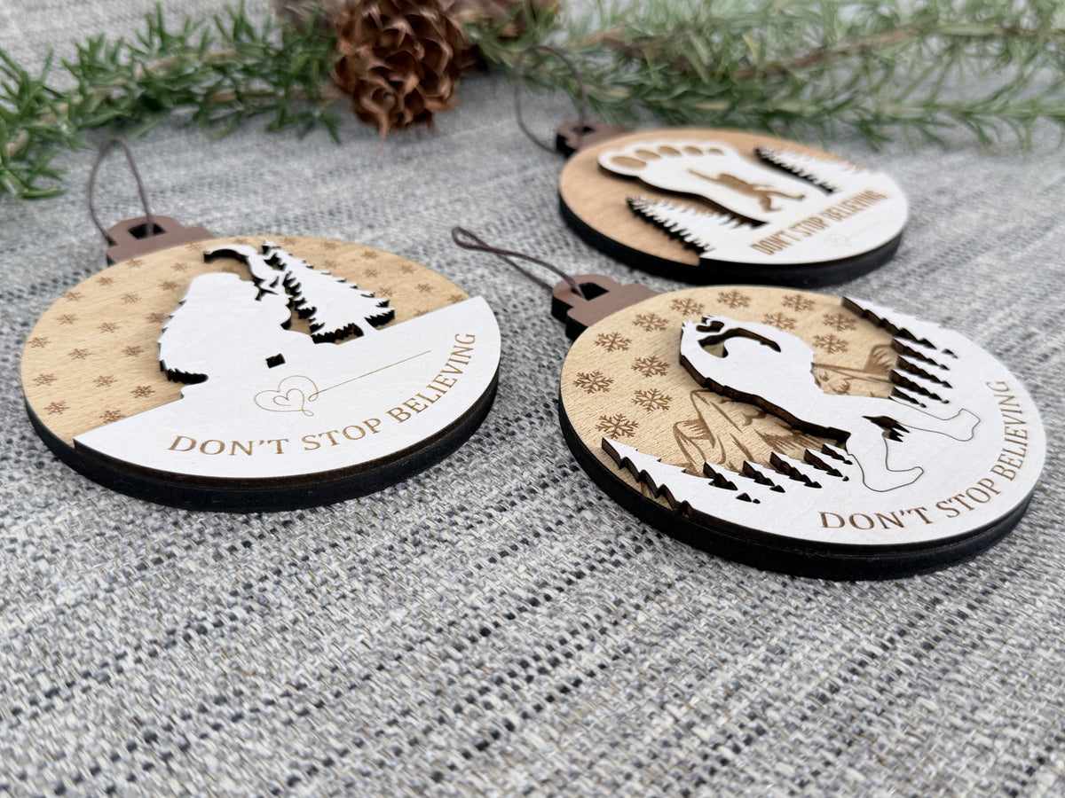 Close-up of three layered wooden Bigfoot Christmas ornaments on textured fabric, highlighting the raised Sasquatch, tree, and mountain designs.