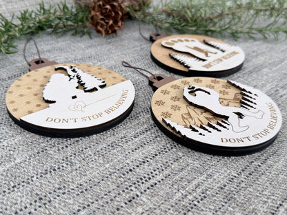 Close-up of three layered wooden Bigfoot Christmas ornaments on textured fabric, highlighting the raised Sasquatch, tree, and mountain designs.