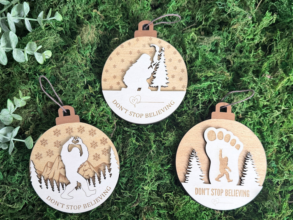 Three layered wooden Sasquatch Christmas ornaments displayed on green moss, featuring Bigfoot silhouettes and Don't Stop Believing engravings.