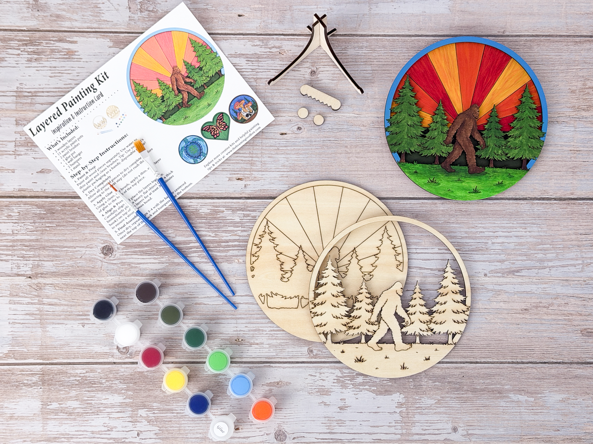 Sasquatch adult craft kit flat lay with unpainted wood layers, finished painted example, paints, brushes, guide, stand pieces, and display hardware.