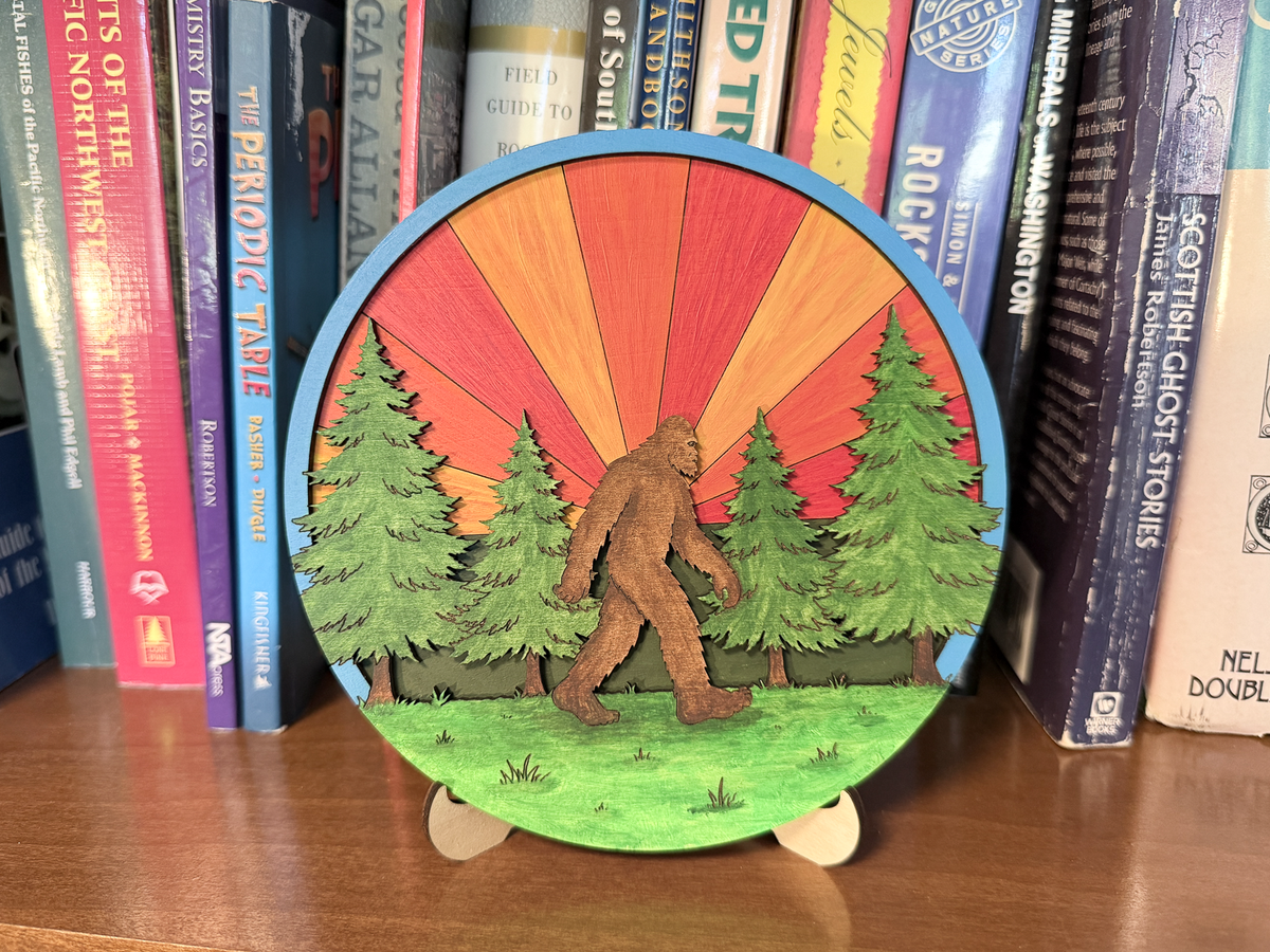 Finished Sasquatch layered wood artwork displayed on a small stand in front of books.