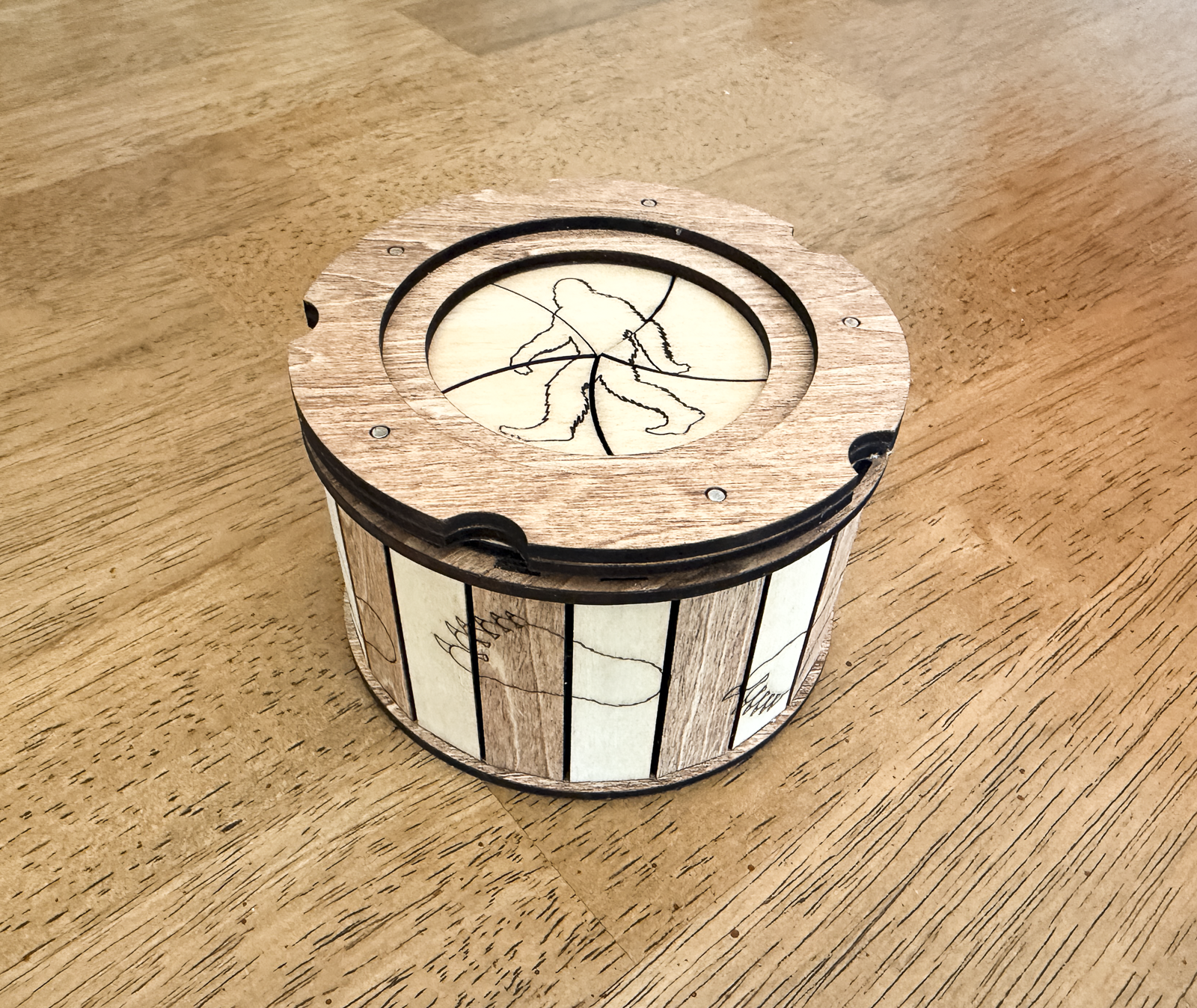 Handcrafted Sasquatch iris box made from basswood with natural wood and mahogany accents, featuring a twist-open lid, Bigfoot silhouette engraving, and footprint details around the sides.