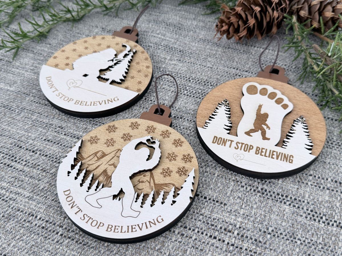 Three handcrafted wooden Sasquatch Christmas ornaments displayed with evergreen branches and pinecones, featuring footprint, mountain, and Christmas tree designs.