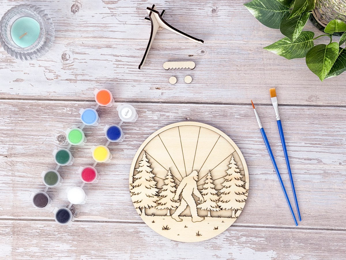 Unpainted Sasquatch adult craft kit arranged with paint pots, brushes, stand pieces, and display hardware on a rustic wood background.