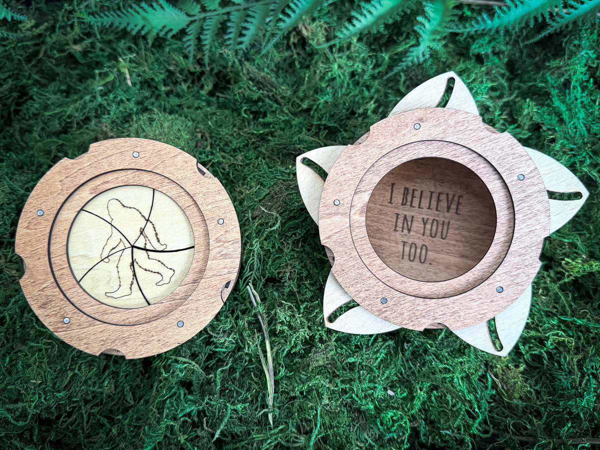 Two round wooden Sasquatch keepsake boxes on green moss, one featuring a Bigfoot silhouette and the other revealing the engraved “I Believe in You Too” message.