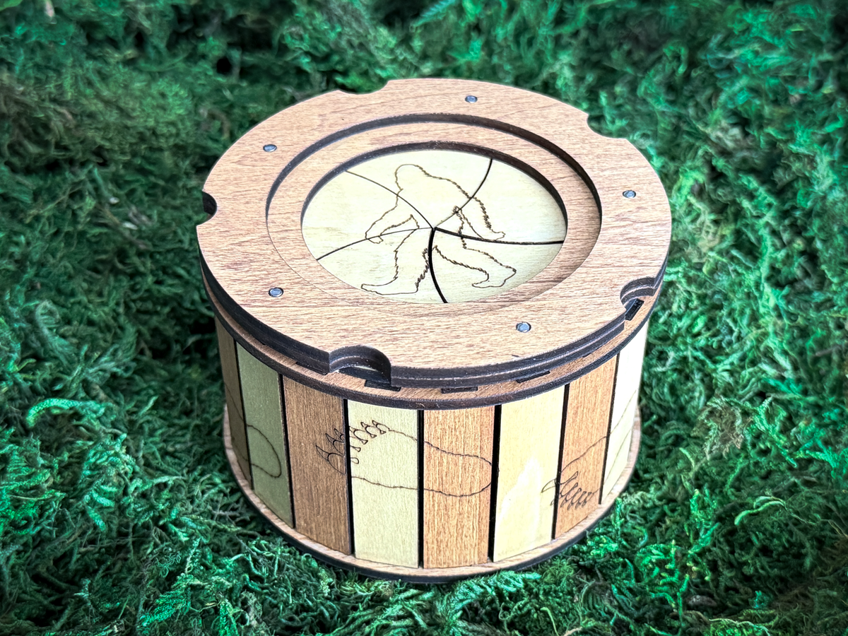 Close-up of a round wooden Sasquatch trinket box with an engraved Bigfoot silhouette, twist-open circular lid, and alternating natural wood panels.