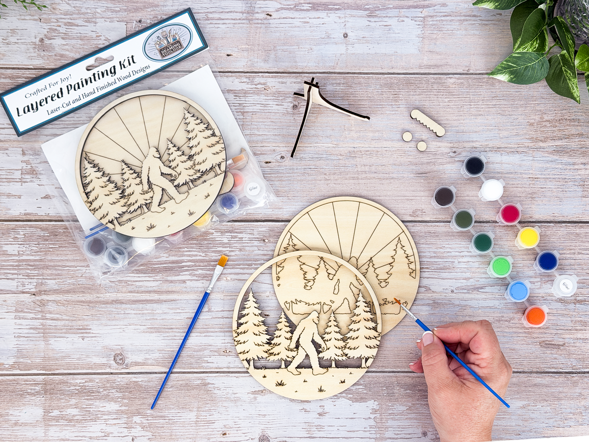 Hand painting the Sasquatch adult craft kit with included brush, paints, wood layers, and packaged kit nearby.