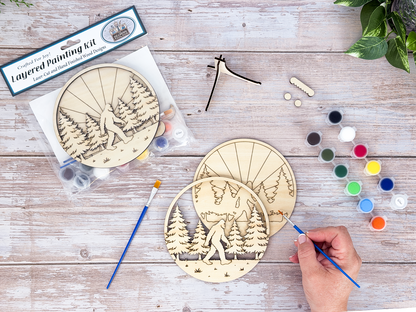 Hand painting the Sasquatch adult craft kit with included brush, paints, wood layers, and packaged kit nearby.