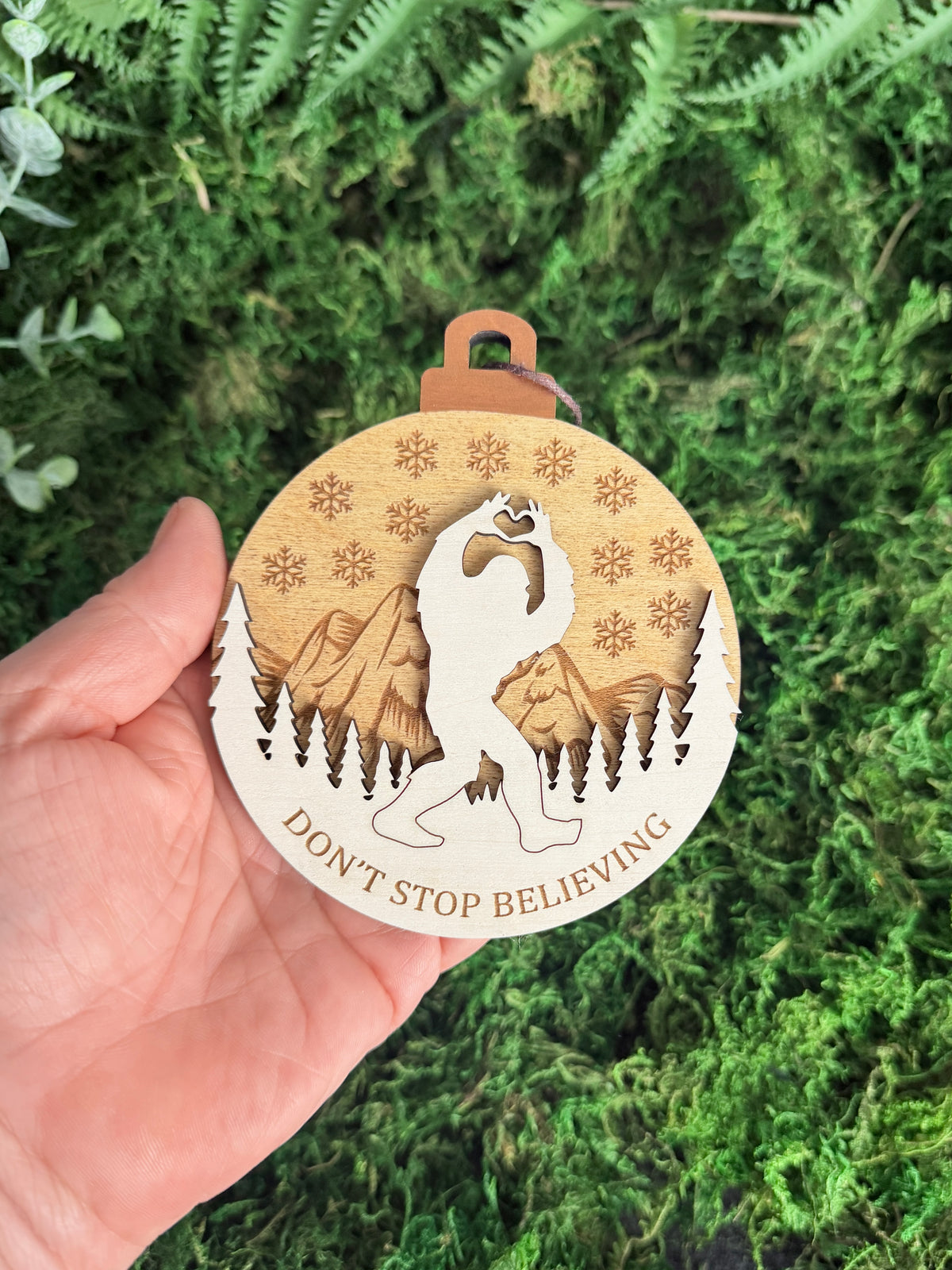 Hand holding a layered wooden Bigfoot Christmas ornament with mountains, pine trees, snowflakes, and Don't Stop Believing engraving to show its size.