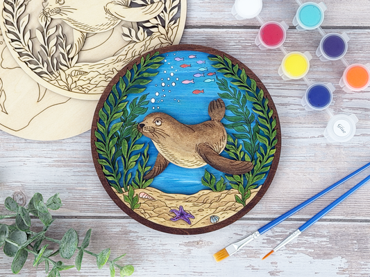 Finished sea lion kelp forest adult craft kit shown with painted layered wood artwork, unpainted kit pieces, paint pots, and brushes on a rustic wood background.
