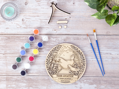 Unpainted Sea Lion custom location wood craft kit laid out with paint pots, brushes, display stand, and hardware on a rustic background.