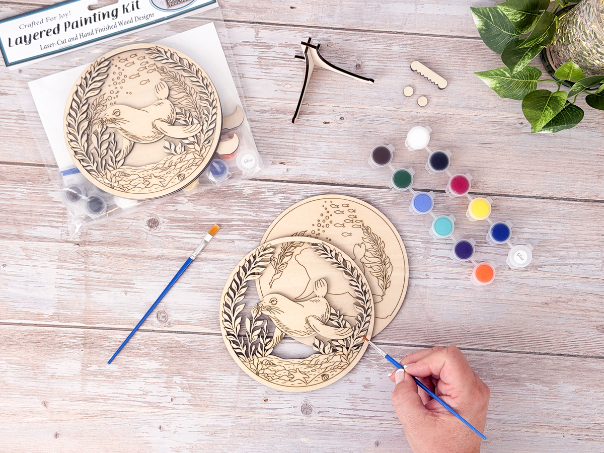 Person painting the Sea Lion layered wood craft kit on a rustic background with paint pots, brushes, packaging, and display hardware.