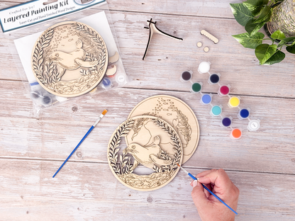 Person painting the Sea Lion layered wood craft kit on a rustic background with paint pots, brushes, packaging, and display hardware.