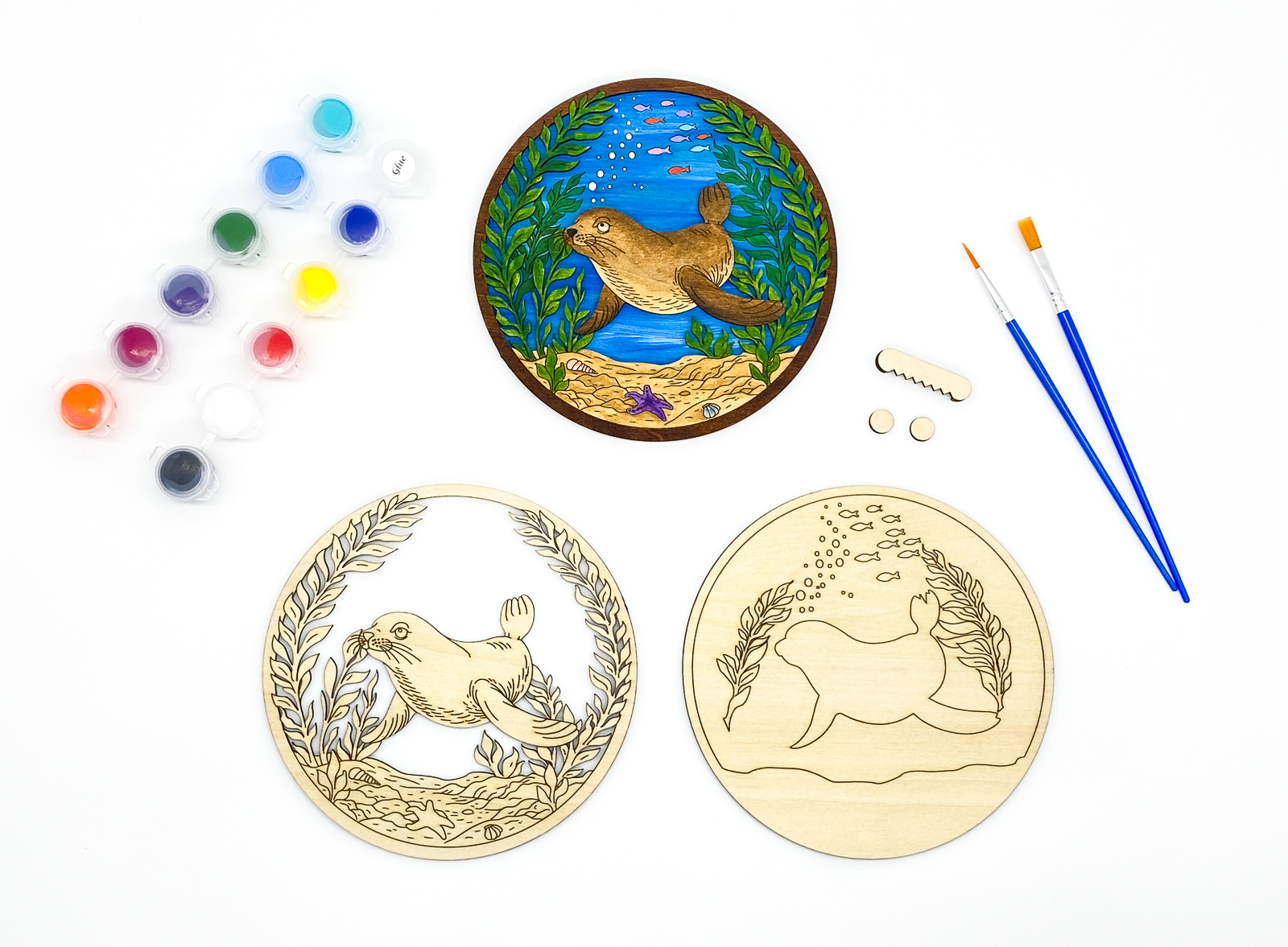 Complete sea lion DIY paint kit with finished piece, unpainted wood layers, paint pots, brushes, hanger and bumpers arranged side by side