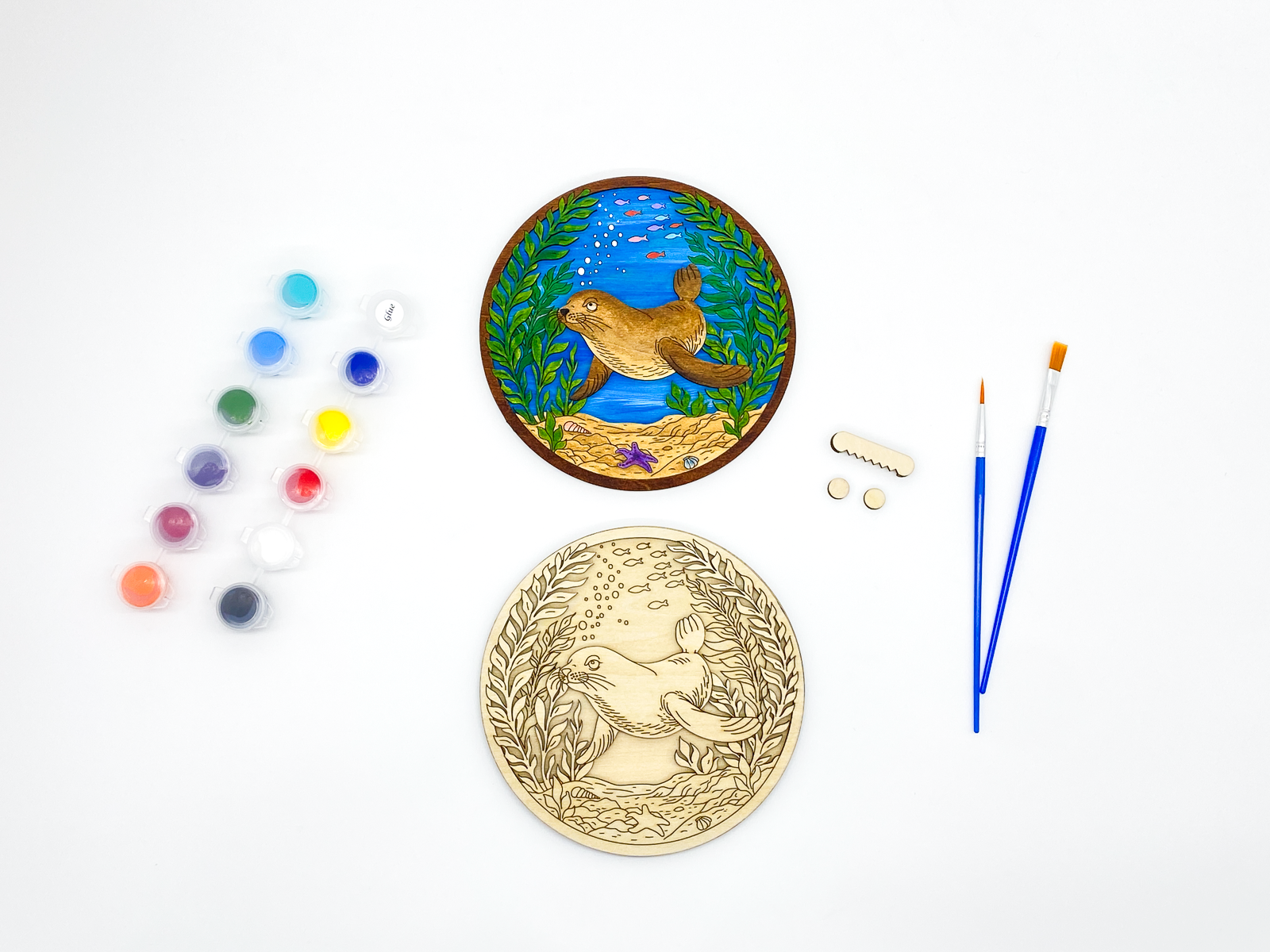 Sea lion kelp forest paint kit with finished sample and stacked wood layers, including paint pots, brushes and hanging hardware