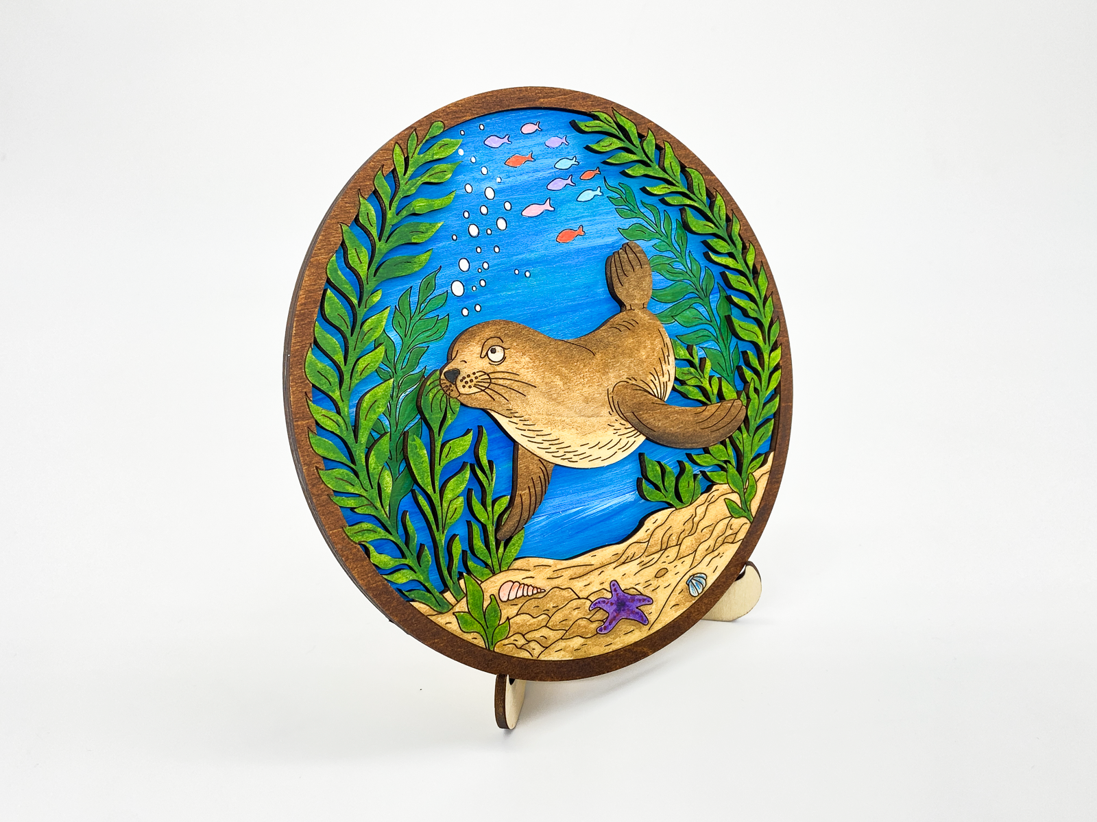 Angled view of painted sea lion kelp forest layered wood art on a white background showing depth and dimension