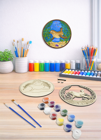 Sea lion kelp forest DIY paint kit on a wood desk with paints, brushes, plant and art supplies, with finished piece displayed on wall