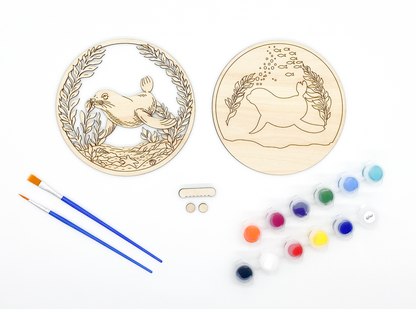 Sea lion DIY paint kit with unpainted wood layers laid flat alongside paint pots, brushes and hanger hardware