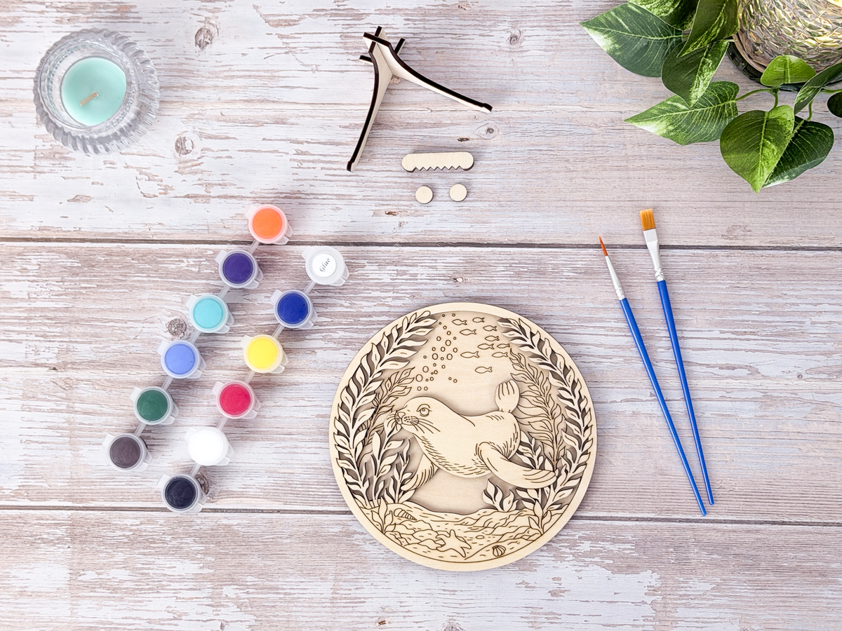 Unpainted Sea Lion layered wood artwork displayed with paint pots, brushes, stand pieces, candle, and greenery on a rustic background.