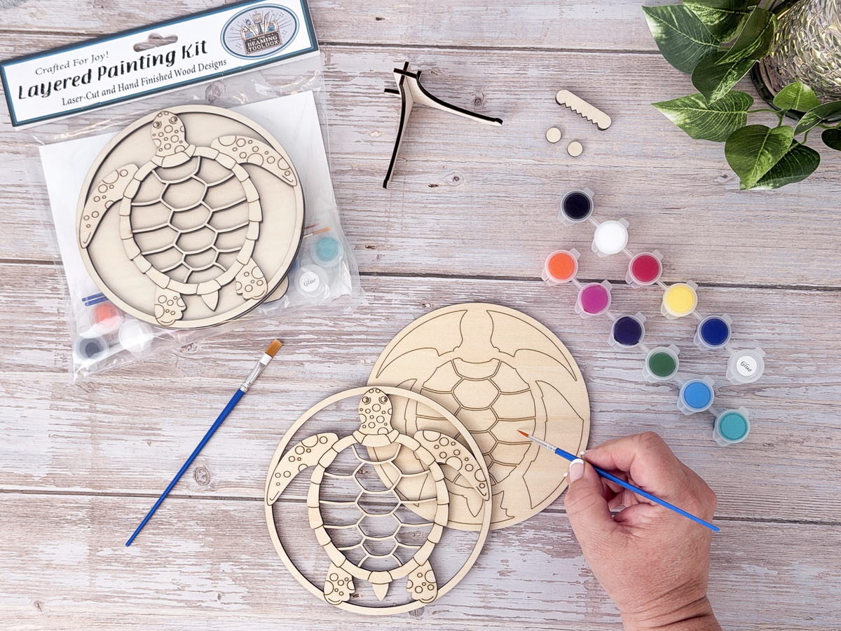 Sea turtle DIY craft kit in progress with a hand painting the wood layer beside paint pots, brushes, packaged kit, and plant decor.