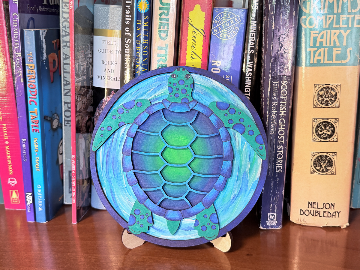 Finished sea turtle layered wood painting displayed on a small stand with blue and green ocean colors in front of books.