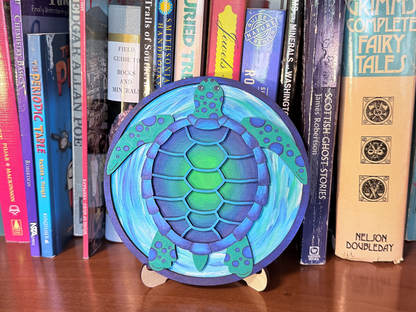 Finished sea turtle layered wood painting displayed on a small stand with blue and green ocean colors in front of books.
