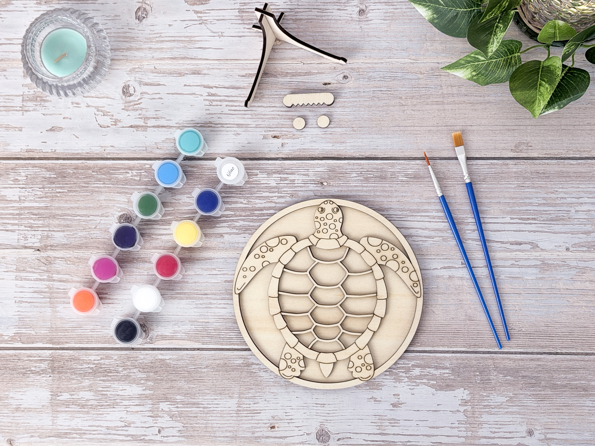 Unpainted sea turtle relaxing craft kit arranged with paint pots, brushes, glue, display stand pieces, candle, and plant decor.