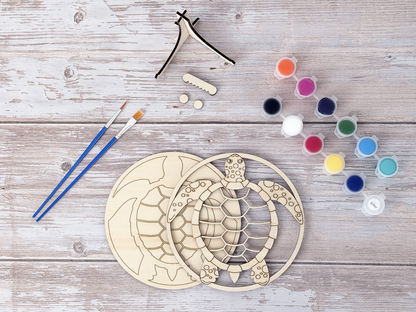Unpainted sea turtle wood craft kit layers arranged with paint pots, blue brushes, glue, and display stand pieces on a rustic wood background.