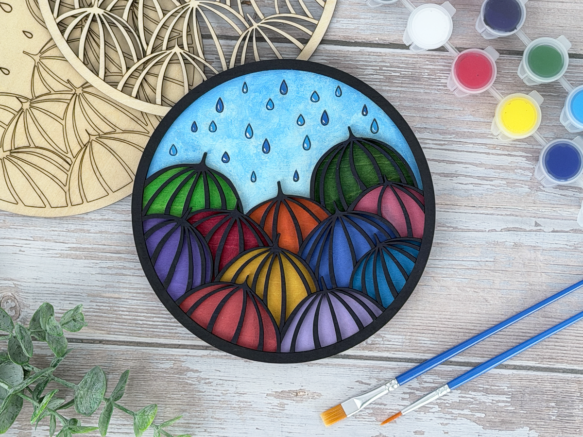 Finished umbrellas adult craft kit shown with painted layered wood artwork, unpainted kit pieces, paint pots, and brushes on a rustic wood background.