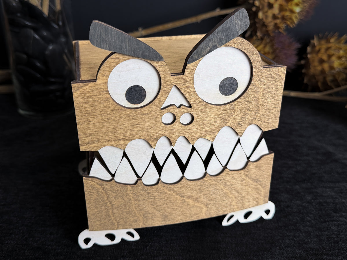 Walnut-stained wooden monster storage box with a closed toothy mouth, large white eyes, black eyebrows, and visible wood grain.