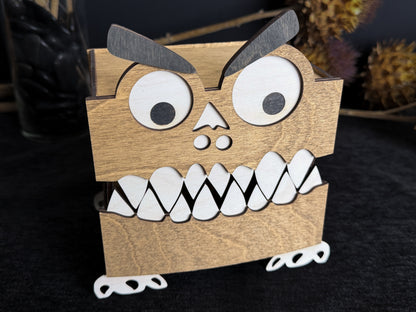 Walnut-stained wooden monster storage box with a closed toothy mouth, large white eyes, black eyebrows, and visible wood grain.