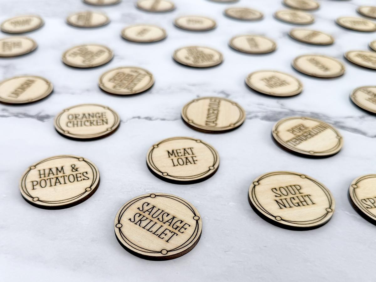 Wooden meal planning coins spread across a marble surface with engraved dinner ideas for choosing weekly meals.