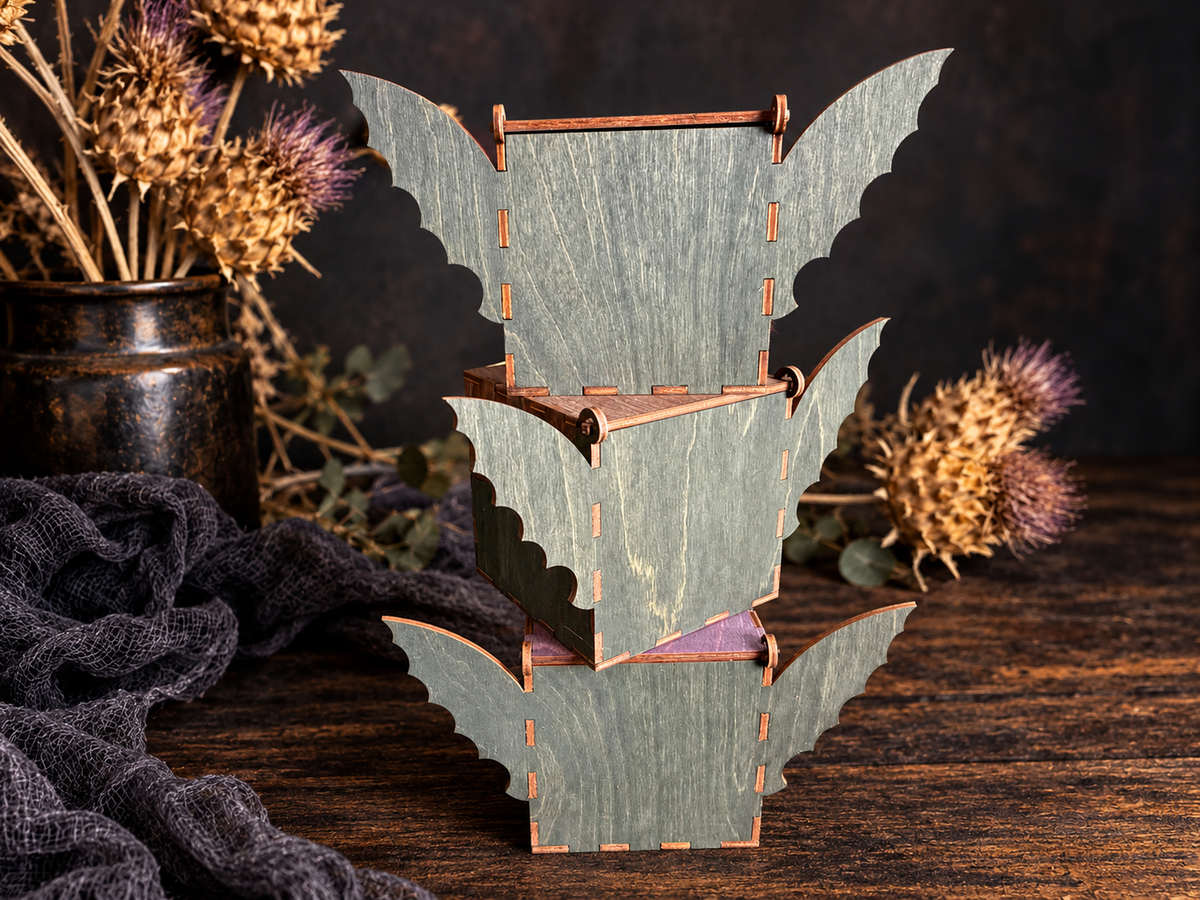 Three handmade wooden bat boxes stacked vertically, showing dark stained bat wings, wood grain, and laser-cut construction against a rustic Halloween backdrop.