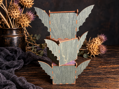 Three handmade wooden bat boxes stacked vertically, showing dark stained bat wings, wood grain, and laser-cut construction against a rustic Halloween backdrop.
