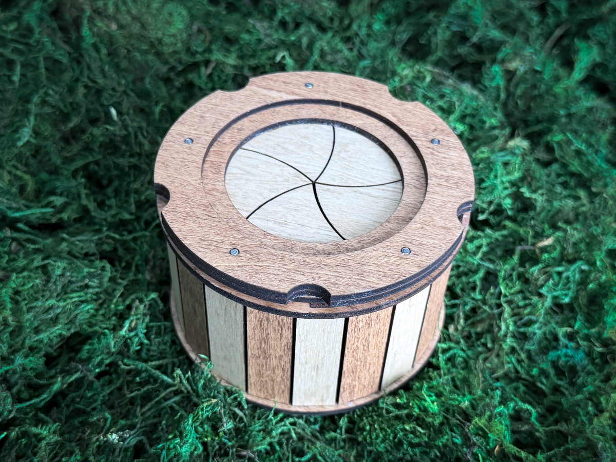 A cylindrical wooden box featuring a mechanical iris lid with curved segments. The exterior walls are constructed from alternating vertical slats of light and dark wood, while the top surface is secured with small metal fasteners.