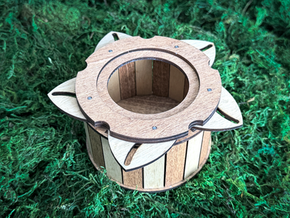 A cylindrical wooden container featuring alternating light and dark wood vertical slats. The top is adorned with a circular wooden lid that has six petal-shaped extensions radiating outward, secured with small metal fasteners.