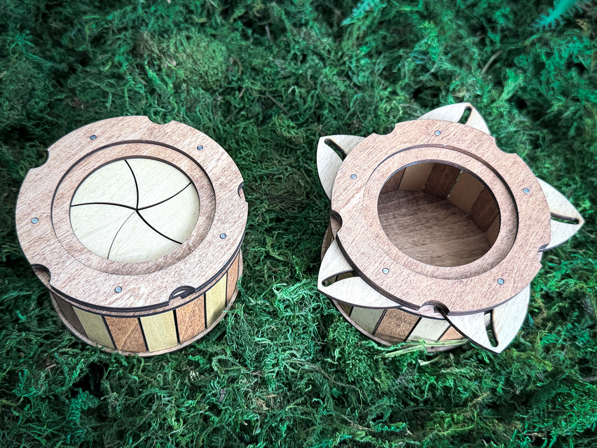 A round wooden box featuring a mechanical iris lid with five curved segments. The sides of the container are constructed from alternating vertical panels of light and dark wood, creating a striped pattern around the cylindrical body.