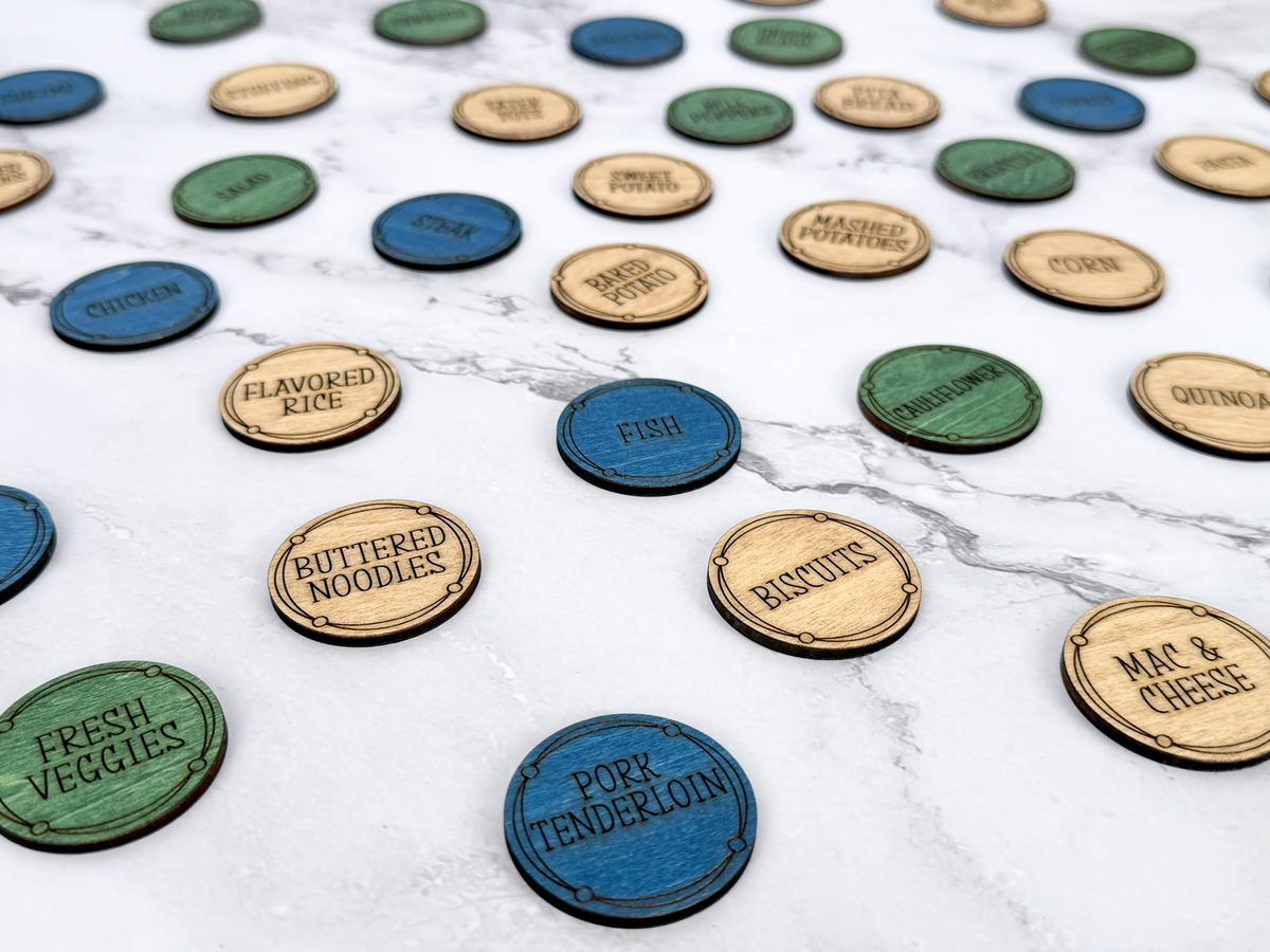 A collection of round, engraved wooden and painted food-themed tokens scattered on a white marble surface. The tokens include natural wood, blue, green, and dark blue colors, with engraved text such as 'Flavored Rice', 'Buttered Noodles', 'Fish', 'Pork Tenderloin', and 'Fresh Veggies'.
