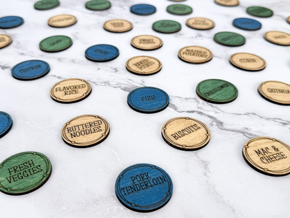 A collection of round, engraved wooden and painted food-themed tokens scattered on a white marble surface. The tokens include natural wood, blue, green, and dark blue colors, with engraved text such as 'Flavored Rice', 'Buttered Noodles', 'Fish', 'Pork Tenderloin', and 'Fresh Veggies'.
