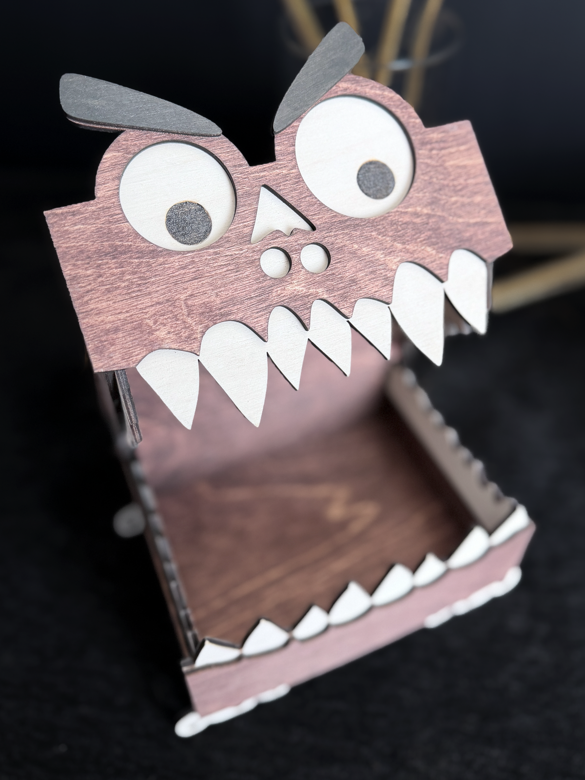 Close-up of an open mahogany-stained wooden monster box with large googly eyes, black eyebrows, white teeth, and a hinged mouth.